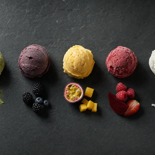 Five scoops of gelato in a row on charcoal slate, each with its own raw ingredient beside it.