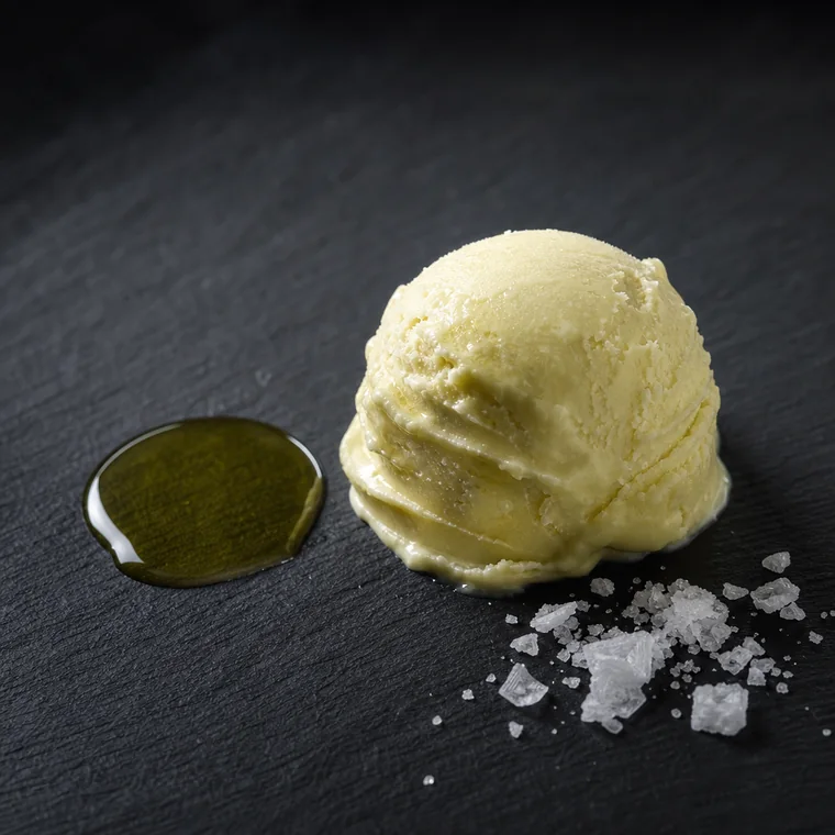 A pale cream scoop of olive oil gelato beside a shallow pool of golden-green olive oil and a scattering of flaky sea salt.