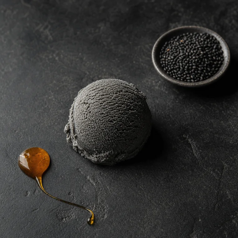 A scoop of dark slate-grey black sesame gelato beside a dish of glossy black sesame seeds and a thread of amber honey.