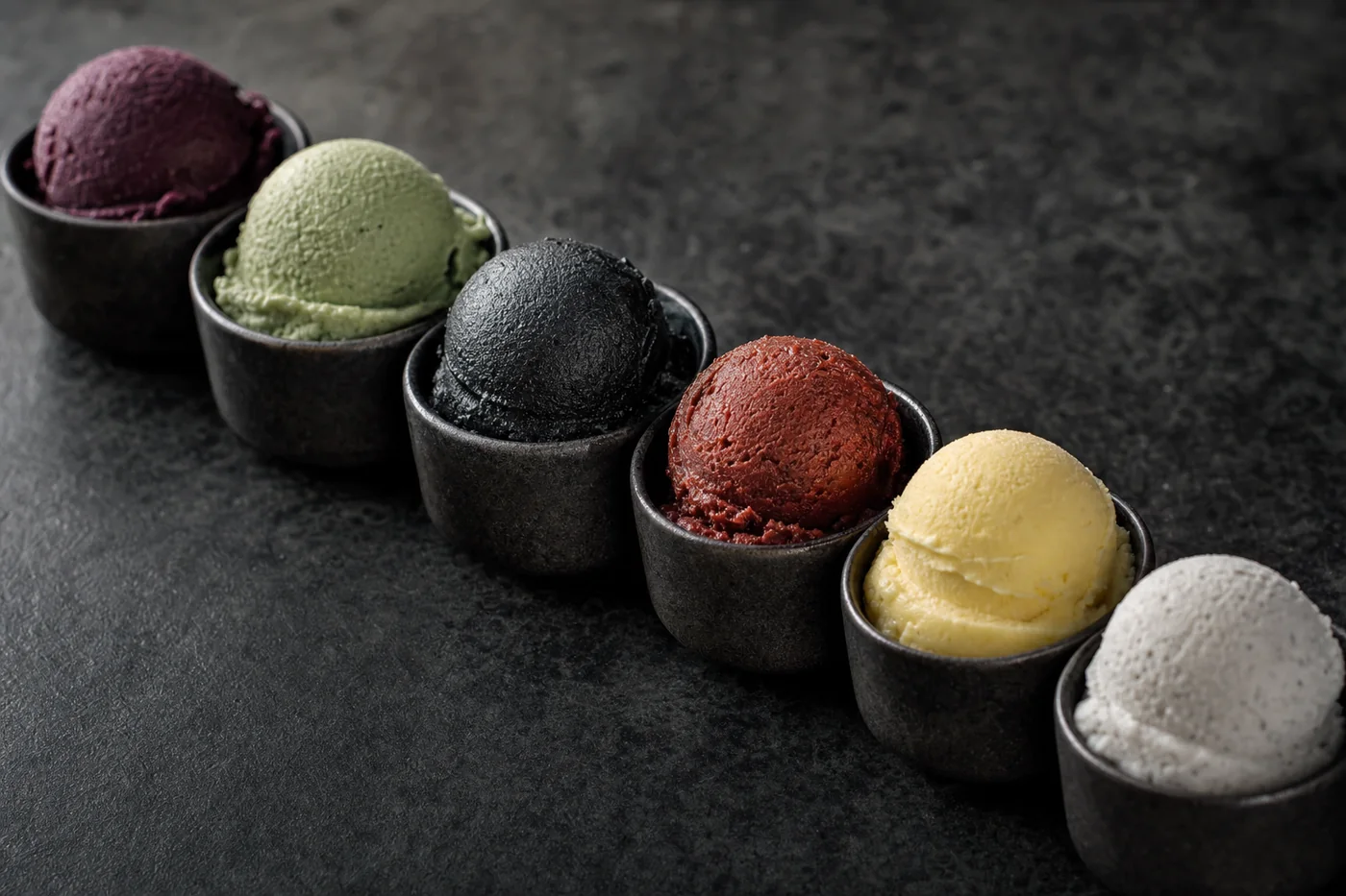 Six small dark cups on charcoal stone, each holding a scoop of a different gelato: violet fig, sage basil, black sesame, chilli chocolate, golden olive oil and pale sea salt.