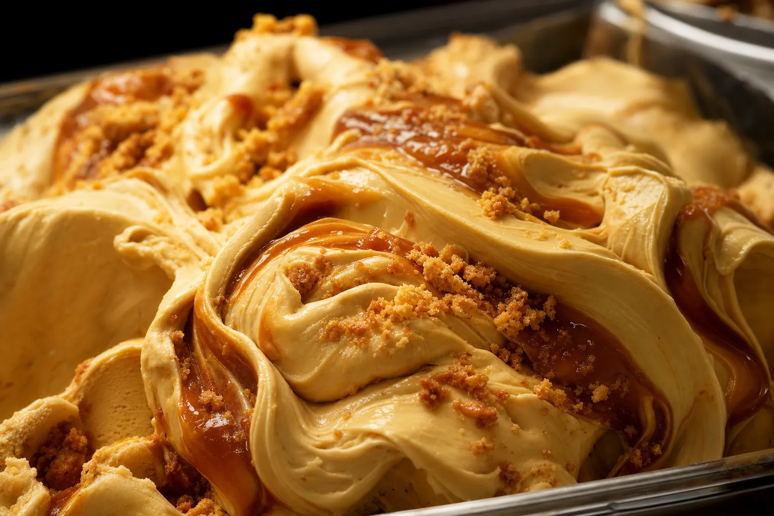 A steel pan of dulce de leche gelato on a gelateria counter, its surface folded into caramel swirls with crumbs of caramelised milk over it.